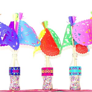 May include: Six glass bottles filled with colourful confetti and topped with paper flags. The flags are cut in a delicate pattern and are in shades of yellow, purple, blue, red, green, and pink.