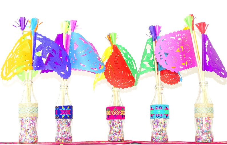May include: Six glass bottles filled with colourful confetti and topped with paper flags. The flags are cut in a delicate pattern and are in shades of yellow, purple, blue, red, green, and pink.