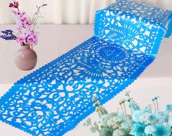 May include: A vibrant blue table runner with intricate floral cut-out designs. The runner is displayed on a white surface, with a vase of pink and purple flowers on the left and a bouquet of blue flowers on the right.