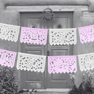 May include: A string of pink and white paper banners with cut-out designs, hanging in front of a doorway. The banners feature various designs, including hearts, stars, and other festive motifs.