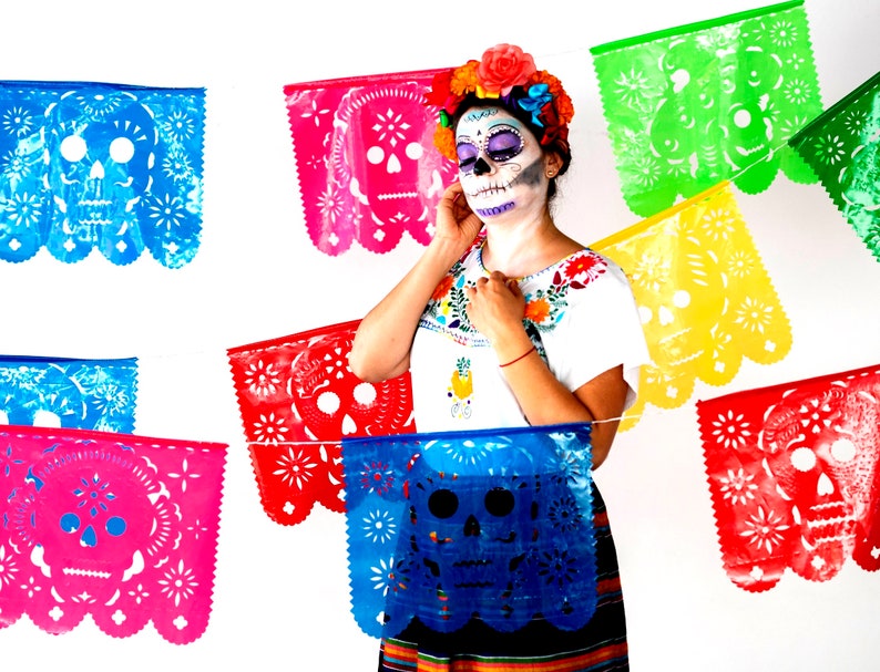May include: A woman wearing traditional Day of the Dead makeup with colorful paper decorations in the background. The decorations feature a skull design and are strung together in a festive garland.