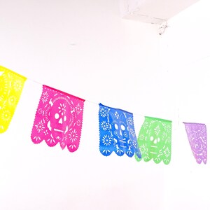 May include: A colorful paper banner with cut-out skull designs in yellow, pink, blue, green, purple, and orange. The banner is strung up against a white wall.