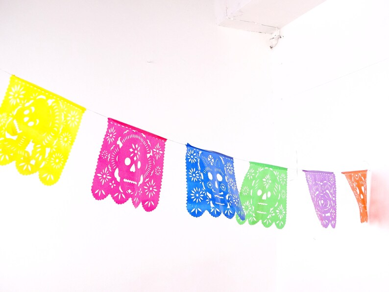 May include: A colorful paper banner with cut-out skull designs in yellow, pink, blue, green, purple, and orange. The banner is strung up against a white wall.