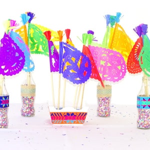 May include: A collection of colourful paper flags with intricate cut-outs, arranged in a vase filled with confetti. The flags are in shades of purple, pink, red, green, yellow, and blue. The vase is a clear glass bottle with a decorative band around the middle.