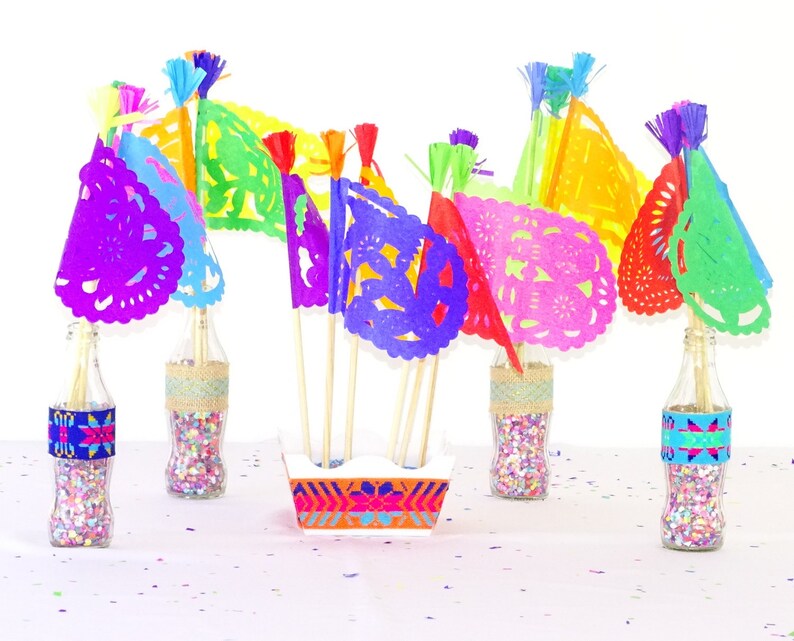 May include: A collection of colourful paper flags with intricate cut-outs, arranged in a vase filled with confetti. The flags are in shades of purple, pink, red, green, yellow, and blue. The vase is a clear glass bottle with a decorative band around the middle.