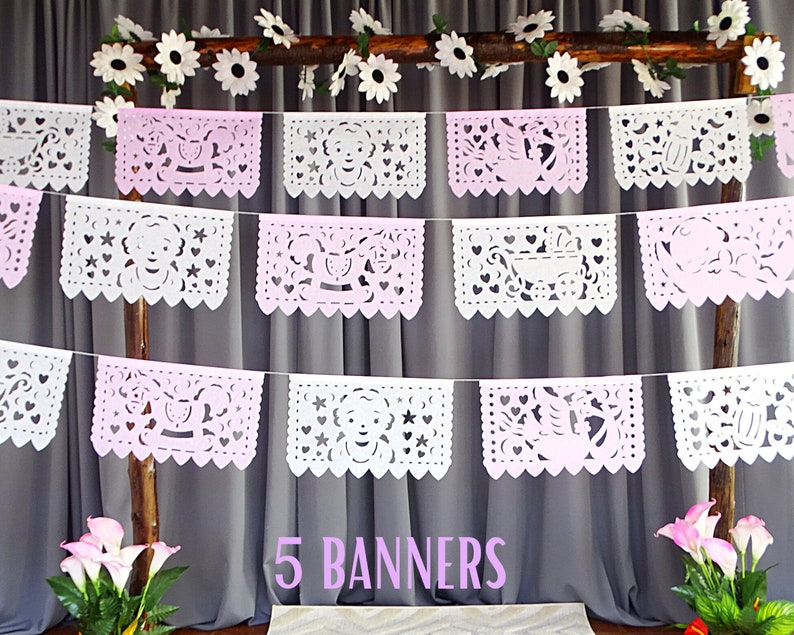 May include: Five white paper banners with pink accents and cut-out designs hang from a wooden branch. The banners feature various baby-themed designs, including a carriage, a star, and a heart. The text "5 BANNERS" is printed below the banners.
