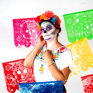 May include: A woman wearing a white blouse with colorful embroidery and a sugar skull makeup design. She is wearing a flower crown and is standing in front of a colorful paper banner with cutouts of skulls.