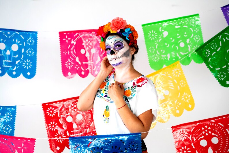 May include: A woman wearing a white blouse with colorful embroidery and a sugar skull makeup design. She is wearing a flower crown and is standing in front of a colorful paper banner with cutouts of skulls.