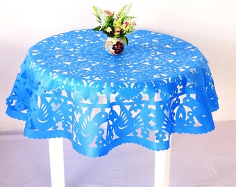 May include: A round, bright blue table cloth with intricate cut-out designs of birds, flowers, and geometric patterns. The cloth is draped over a white table. A small vase with colorful flowers sits on top.