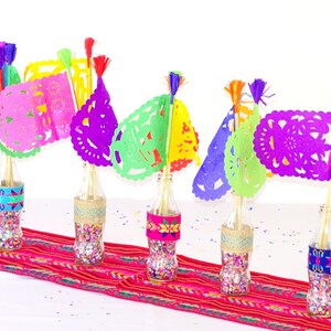 May include: Five glass bottles filled with colourful confetti and topped with colourful paper cutouts, resembling flags, with tassels. The bottles are arranged on a red and white striped table runner.