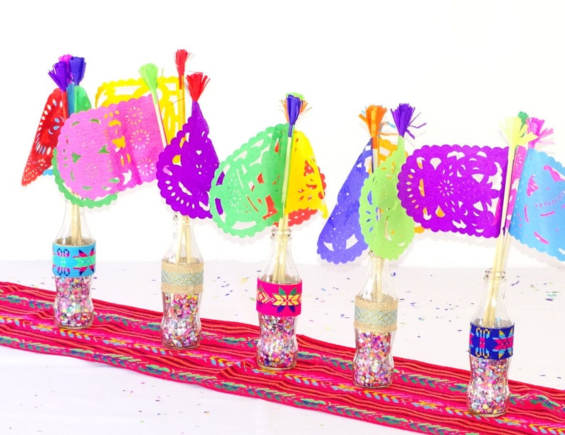 May include: Five glass bottles filled with colourful confetti and topped with colourful paper cutouts, resembling flags, with tassels. The bottles are arranged on a red and white striped table runner.