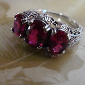 May include: A silver ring featuring three oval, deep red gemstones. The ring has an ornate, filigree design around the gemstones and along the band. The gemstones are set close together, creating a bold and eye-catching piece of jewelry.