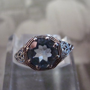 May include: A silver ring with an octagonal, light blue gemstone. The ring has an ornate, filigree design around the gemstone and on the band. The ring is set on a clear, glass-like surface.