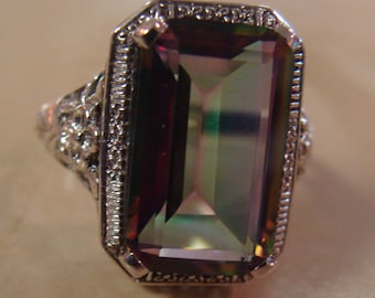 May include: A silver ring featuring a large, rectangular gemstone with a color-shifting effect, displaying hues of purple, green, and gold. The ring has an ornate, filigree design on the band and around the gemstone setting.