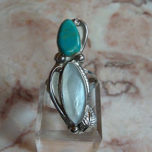 May include: A silver ring featuring a teardrop-shaped turquoise stone and a large, oval, pearlescent stone. The ring has a decorative, leaf-like design on the sides and is displayed on a clear acrylic stand.