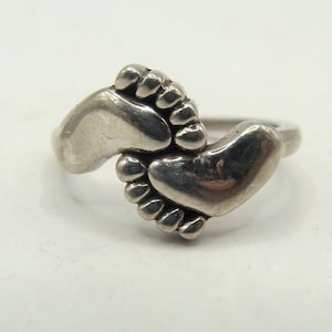 May include: A silver ring featuring a design of two baby footprints. The ring is made of a shiny, metallic material and is set against a white background. The footprints are detailed and appear to be three-dimensional.