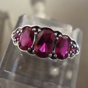 May include: A silver ring featuring three prominent oval-cut deep pinkish-red stones arranged horizontally, flanked by two smaller round stones on each side, all set within a decorative metal frame with small bead-like accents along the edges.