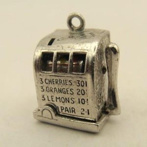 May include: A silver-toned metal charm shaped like a miniature slot machine. The front features engraved text reading 3 CHERRIES 30, 3 ORANGES 20, 3 LEMONS 10, and 1 PAIR 2. It includes a side lever and a small circular loop at the top for attachment.