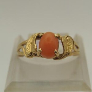 Op de afbeelding: Een gouden ring met een ovale, oranjerode edelsteen. De band van de ring heeft aan weerszijden van de edelsteen een bladvormig ontwerp. De ring wordt gepresenteerd op een heldere, schuine standaard tegen een witte achtergrond.