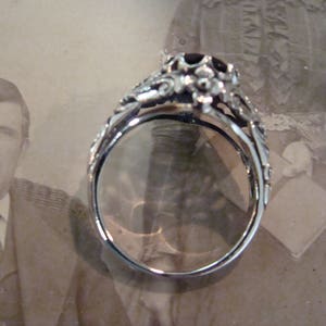 May include: A silver ring with an ornate floral design and a dark gemstone at the center. The ring's band is detailed with delicate scrollwork. The ring is set against a vintage photograph background.