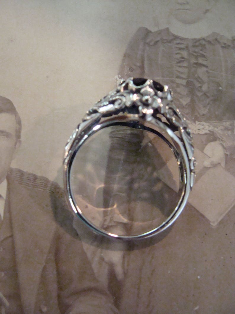 May include: A silver ring with an ornate floral design and a dark gemstone at the center. The ring's band is detailed with delicate scrollwork. The ring is set against a vintage photograph background.