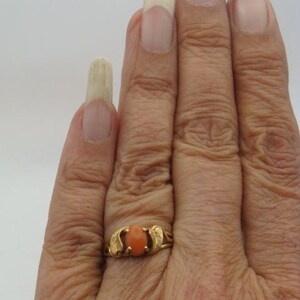 Op de afbeelding: Een gouden ring met een ovale, oranje steen. De ring heeft een delicaat ontwerp met bladvormige accenten aan weerszijden van de steen. De ring zit om een vinger.