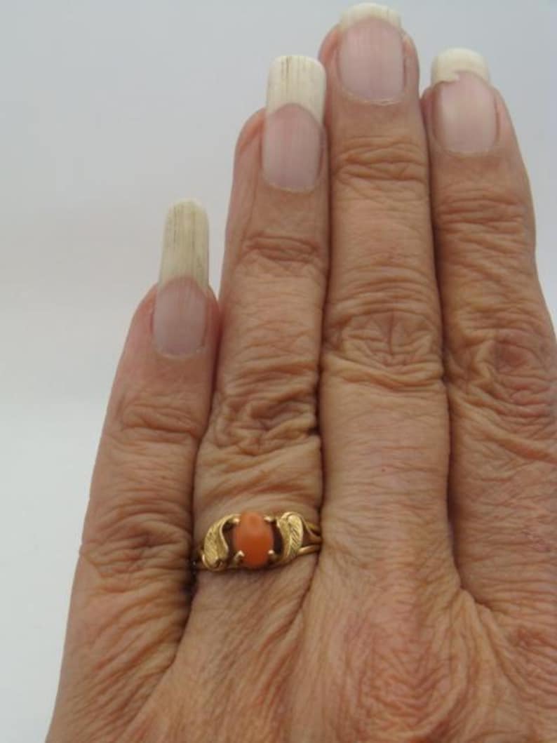 Op de afbeelding: Een gouden ring met een ovale, oranje steen. De ring heeft een delicaat ontwerp met bladvormige accenten aan weerszijden van de steen. De ring zit om een vinger.