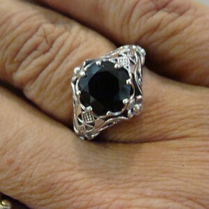 May include: A silver ring featuring a large, faceted black gemstone. The ring has an ornate, filigree design with floral and scroll details. The gemstone is set in a square shape, and the ring is worn on a finger.