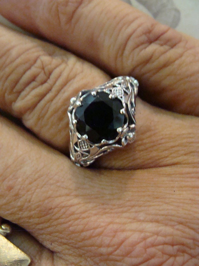 May include: A silver ring featuring a large, faceted black gemstone. The ring has an ornate, filigree design with floral and scroll details. The gemstone is set in a square shape, and the ring is worn on a finger.