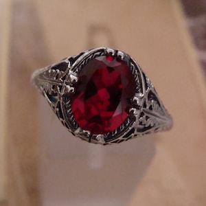 May include: A silver ring with an oval, deep red gemstone. The ring features an ornate, filigree design around the gemstone and along the band. The gemstone is the focal point, set in a detailed, vintage-style setting.