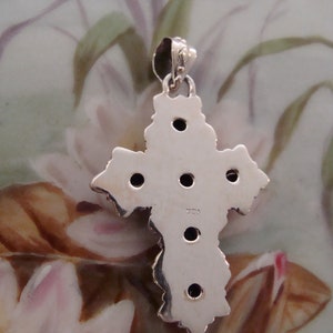 Puede incluir: Un colgante de plata con forma de cruz, con un dise&ntilde;o texturizado y peque&ntilde;as piedras oscuras. El colgante tiene un lazo para una cadena. La parte posterior del colgante es visible, mostrando el sello 925, que indica la pureza del metal.