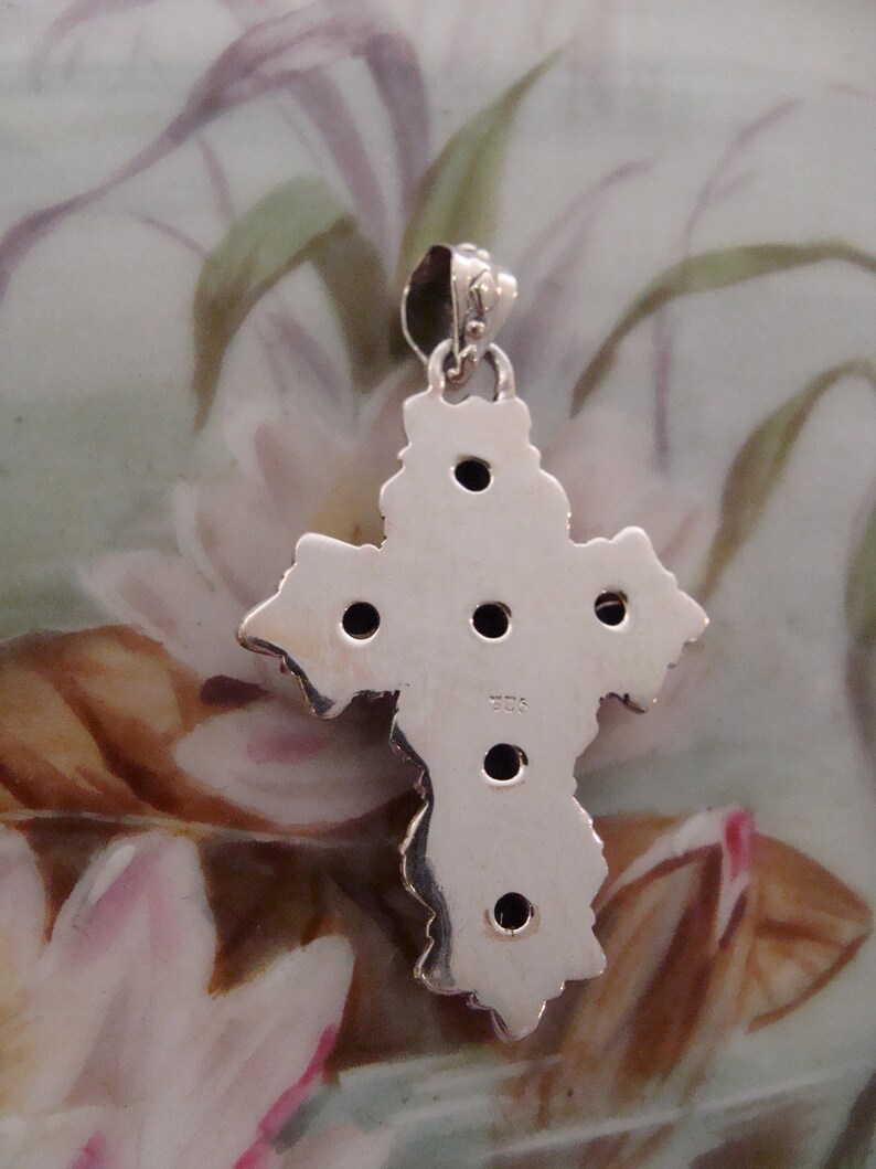 Puede incluir: Un colgante de plata con forma de cruz, con un dise&ntilde;o texturizado y peque&ntilde;as piedras oscuras. El colgante tiene un lazo para una cadena. La parte posterior del colgante es visible, mostrando el sello 925, que indica la pureza del metal.