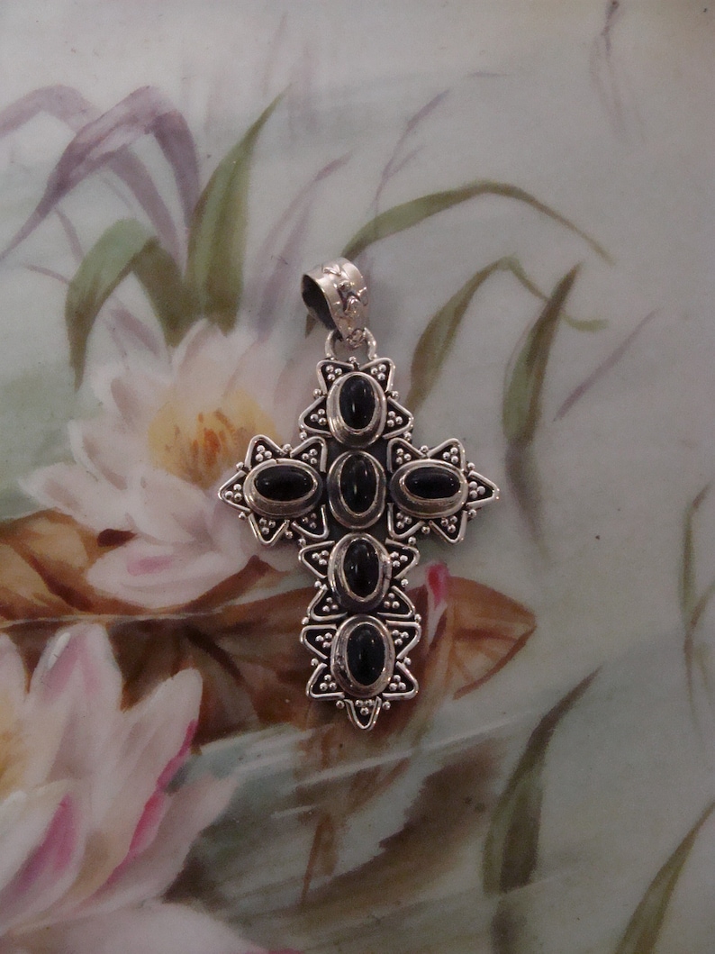 Puede incluir: Un colgante de cruz plateado con piedras negras ovaladas. La cruz tiene un dise&ntilde;o detallado con peque&ntilde;os detalles de cuentas y una fianza decorativa. El colgante probablemente est&aacute; hecho de plata de ley y es un accesorio religioso o de moda.