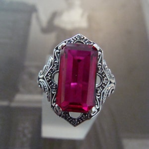 May include: A silver ring with a large, rectangular, magenta-colored gemstone. The ring has an ornate, filigree design around the gemstone and on the band. The ring is displayed against a blurred background.