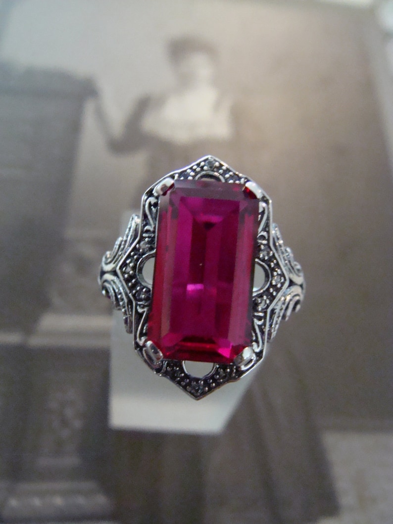 May include: A silver ring with a large, rectangular, magenta-colored gemstone. The ring has an ornate, filigree design around the gemstone and on the band. The ring is displayed against a blurred background.