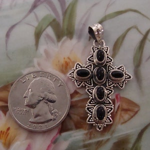 Puede incluir: Colgante de cruz plateada con piedras negras ovaladas. Una moneda est&aacute; al lado para comparar el tama&ntilde;o. El colgante tiene un dise&ntilde;o detallado con detalles en forma de estrella, probablemente una joya.