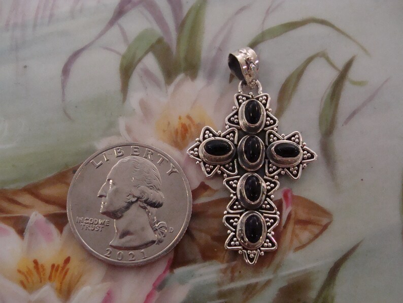 Puede incluir: Colgante de cruz plateada con piedras negras ovaladas. Una moneda est&aacute; al lado para comparar el tama&ntilde;o. El colgante tiene un dise&ntilde;o detallado con detalles en forma de estrella, probablemente una joya.