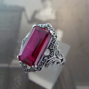 May include: A silver-toned ring featuring a large, rectangular, deep red gemstone. The ring has an ornate, filigree design around the stone and on the band, with small accent stones. The ring is displayed against a blurred background.
