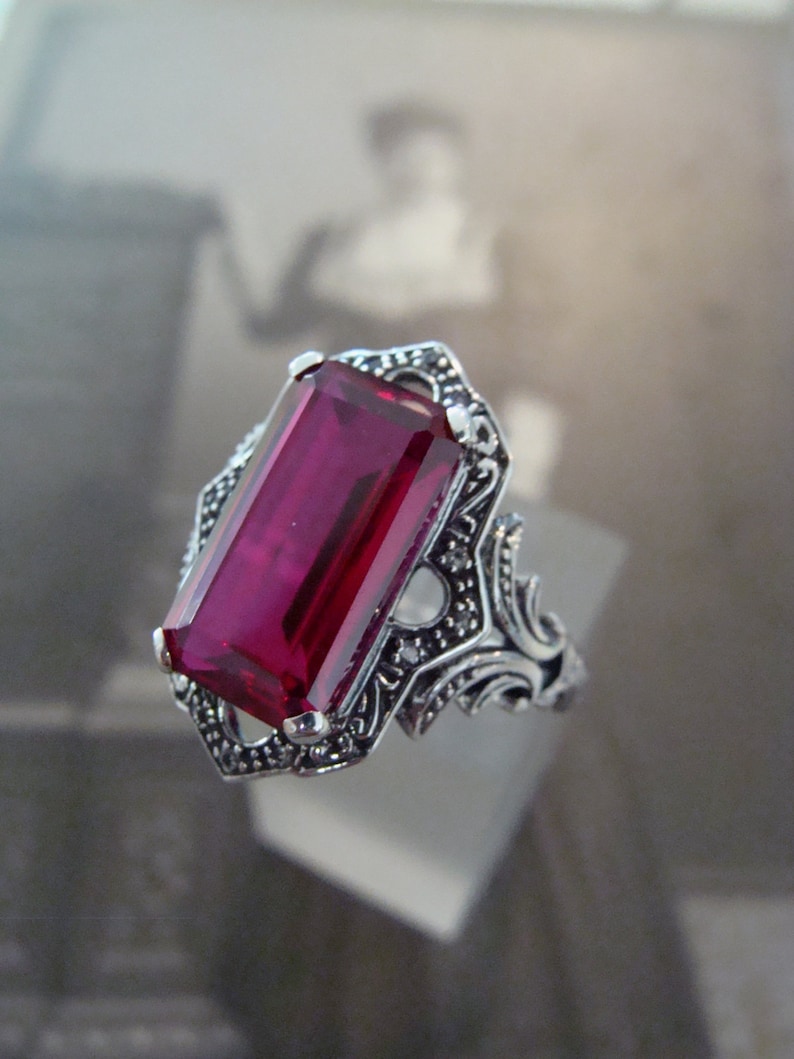 May include: A silver-toned ring featuring a large, rectangular, deep red gemstone. The ring has an ornate, filigree design around the stone and on the band, with small accent stones. The ring is displayed against a blurred background.