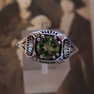 May include: A silver ring with an oval, green gemstone. The ring has an ornate, filigree design around the gemstone and along the band. The gemstone is a vibrant green color, and the ring appears to be vintage or antique.