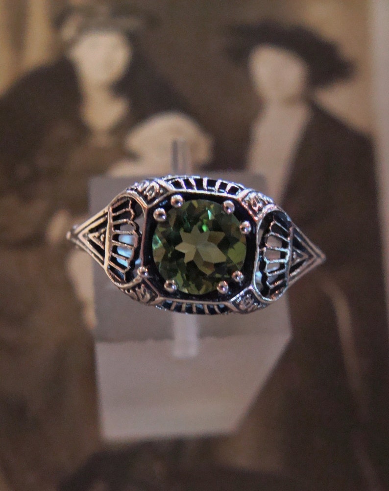 May include: A silver ring with an oval, green gemstone. The ring has an ornate, filigree design around the gemstone and along the band. The gemstone is a vibrant green color, and the ring appears to be vintage or antique.