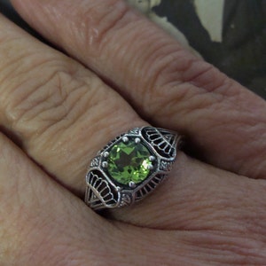 May include: A silver ring with a square, light green gemstone. The ring has an ornate, filigree design around the stone and on the band. The ring is on a person's finger.