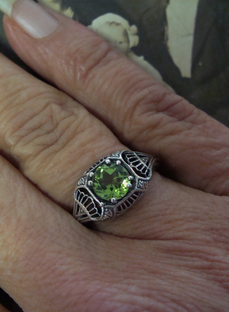 May include: A silver ring with a square, light green gemstone. The ring has an ornate, filigree design around the stone and on the band. The ring is on a person's finger.