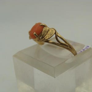 Op de afbeelding: Een gouden ring met een ovale, oranjerode edelsteen. De band van de ring heeft een bladerontwerp aan de zijkanten en de edelsteen wordt op zijn plaats gehouden door vier tanden. De ring wordt tentoongesteld op een heldere, rechthoekige standaard.