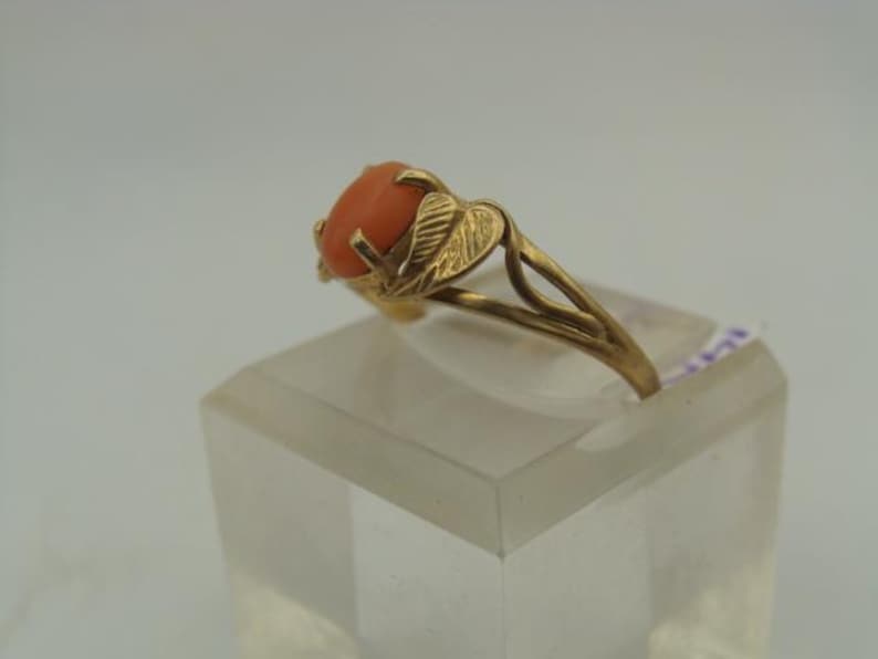 Op de afbeelding: Een gouden ring met een ovale, oranjerode edelsteen. De band van de ring heeft een bladerontwerp aan de zijkanten en de edelsteen wordt op zijn plaats gehouden door vier tanden. De ring wordt tentoongesteld op een heldere, rechthoekige standaard.