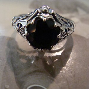 May include: A silver-toned ring with an oval, black gemstone. The ring features intricate filigree detailing around the band and the setting of the stone. The design includes floral and leaf motifs, creating a vintage aesthetic.