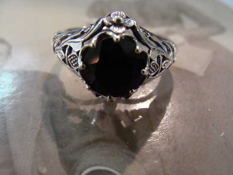 May include: A silver-toned ring with an oval, black gemstone. The ring features intricate filigree detailing around the band and the setting of the stone. The design includes floral and leaf motifs, creating a vintage aesthetic.
