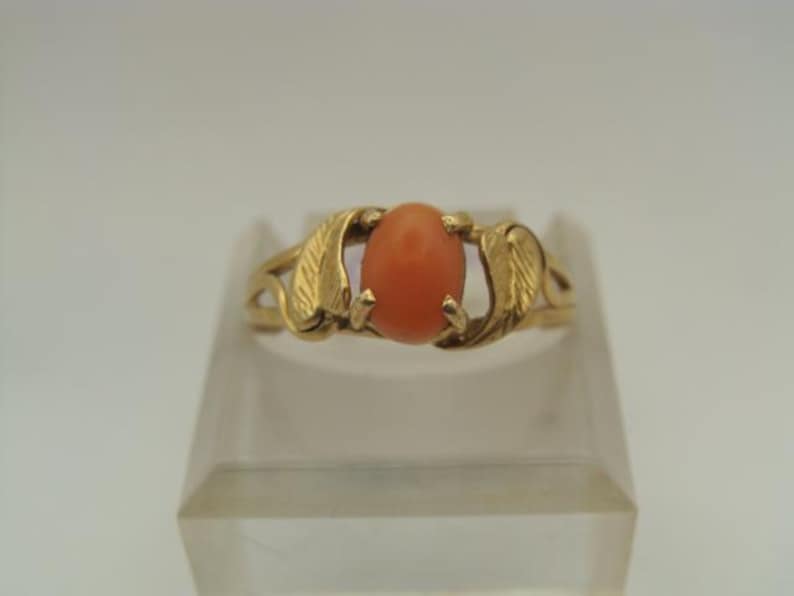 Op de afbeelding: Een gouden ring met een ovale, oranjerode edelsteen. De band van de ring heeft aan weerszijden van de edelsteen een bladvormig ontwerp. De ring wordt gepresenteerd op een heldere, schuine standaard tegen een witte achtergrond.