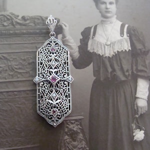 May include: A silver filigree pendant with a rectangular shape, featuring intricate openwork and adorned with small red and white gemstones. The pendant is suspended from a delicate chain. The background is a vintage photograph of a woman.