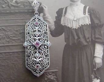 Puede incluir: Un colgante de filigrana plateada con forma rectangular, con intrincados calados y adornado con pequeñas piedras preciosas rojas y blancas. El colgante está suspendido de una delicada cadena. El fondo es una fotografía antigua de una mujer.